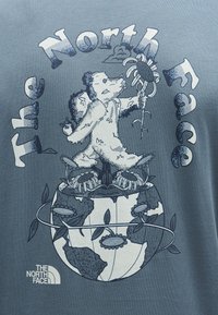 Αρκούδα που κρατάει ένα λουλούδι, στέκεται πάνω σε έναν κόσμο καλυμμένο με λουλούδια και κληματαριές, περιβαλλόμενη από το κείμενο "The North Face" σε καλλιγραφικά γράμματα.