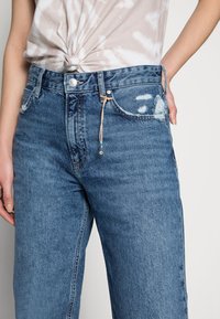 Jeans de tiro alto de color azul con un corte relajado, que presentan detalles desgastados, un cierre de botón en la parte delantera y un charm decorativo de cuentas en la cinturilla.