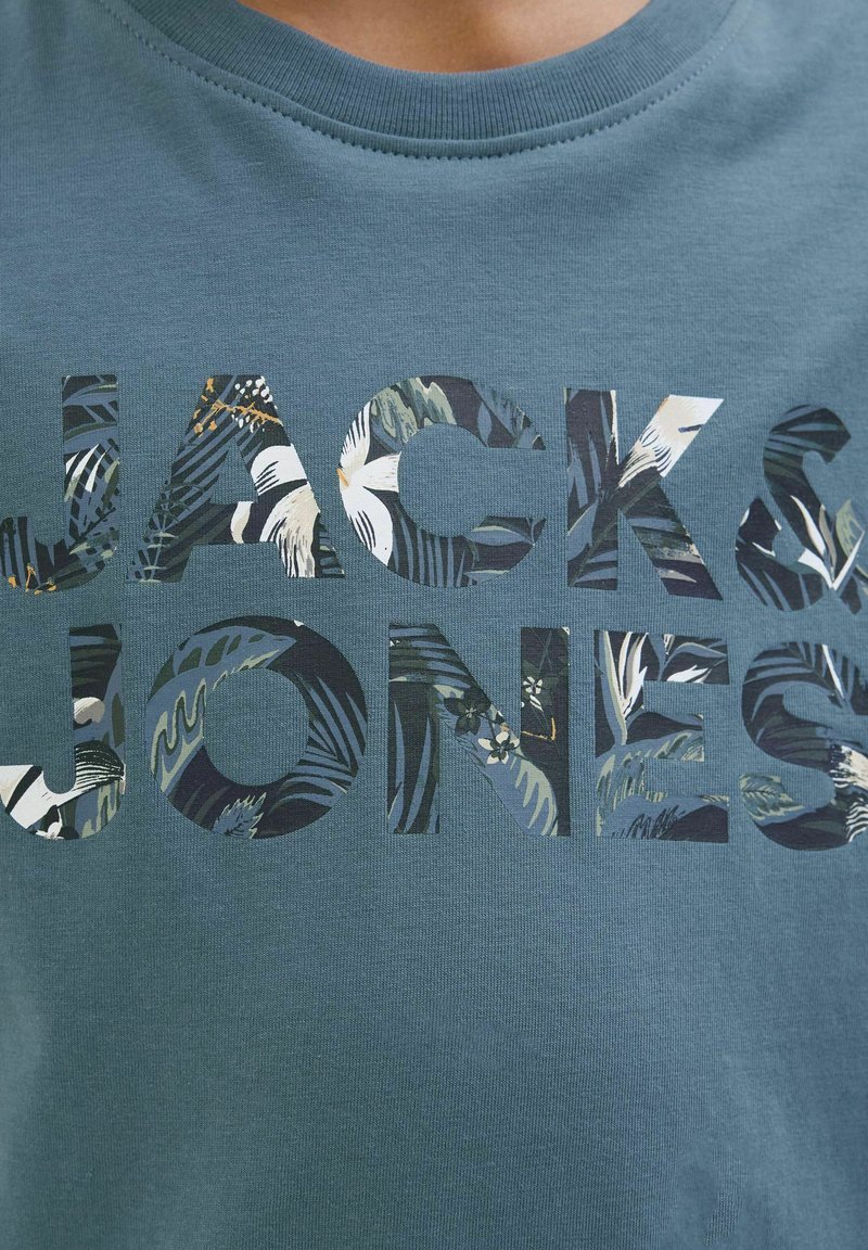 Tirkizna pamučna majica s velikim izrezanim slovima koja tvore natpis "JACK&JONES", ispunjena printom tropskog bilja. Glatka tekstura, okrugli izrez.