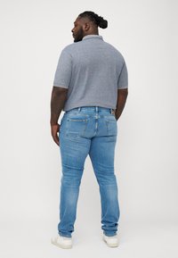 Man med flätat hår som bär en grå polo med kort ärm, ljusblå jeans och vita sneakers, stående vänd bortåt på en vit bakgrund.