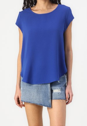 Femme portant un haut bleu uni à manches courtes associé à une minijupe en denim bleu clair asymétrique avec des zones usées.