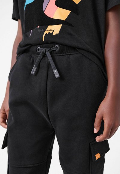 Mustat joggerit, joissa on nauhavyötärö, sivutaskut ja oranssi etiketti. Pehmeä materiaali, suunniteltu mukavuutta ja toimivuutta varten.