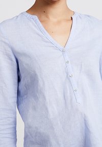 Helles blaues Leinenhemd mit V-Ausschnitt, langen Ärmeln und einer Knopfleiste vorne. Leichter Stoff mit dezenten Farbvariationen.