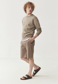 Felpa beige con polsini a costine, abbinata a pantaloni corti beige con tasche laterali. Sandali marroni con due cinturini e dettaglio fibbia.