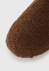Brązowy puszysty kapcie z teksturowanym, miękkim materiałem zewnętrznym i zaokrąglonym czubkiem. Posiada ciemną podeszwę dla zwiększonej trwałości.