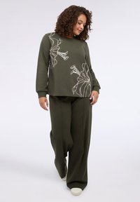 Olivgrön sweatshirt med långa ärmar och blommigt konturdesign, kombinerad med matchande lösa byxor och ljusa skor.