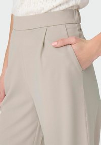 Beżowe spodnie plisowane o gładkiej teksturze, z bocznymi kieszeniami i dopasowanym krojem, z wysokim stanem i bez widocznych akcentów sprzętowych.