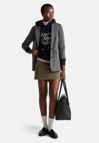 Šedý blazer přes námořnickou mikinu se zlatým textem, světle šedé tričko, olivová sukně, bílé ponožky a černé loafers; drží černou tašku.