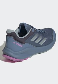 Zapato de senderismo azul oscuro con parte superior de malla, acentos grises y tres franjas reflectantes. Presenta una suela texturizada con un detalle en el talón de color rosa.