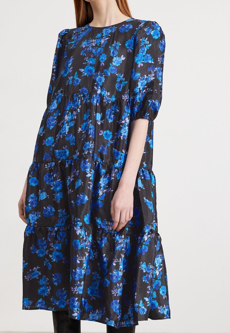 Robe midi noire à étages avec motif floral bleu, manches bouffantes mi-longues aux coudes, et encolure ronde, portée par une femme.