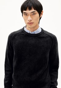 Schwarzer Strickpullover mit geripptem Kragen und Schulternähten, der über einem hellblauen, weich strukturierten Button-up-Hemd getragen wird.