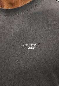 Tumši pelēks kokvilnas T-krekls ar apaļu kakla izgriezumu un baltu drukātu tekstu "Marc O'Polo DENIM" priekšpusē, gluda tekstūra un pieguļošs dizains.