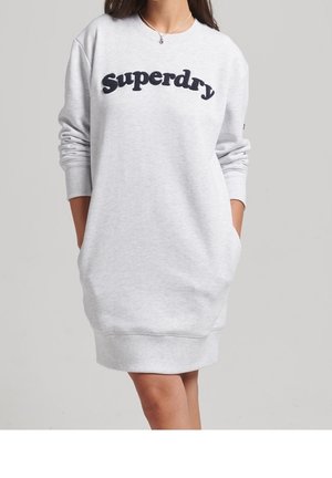Woman wearing a light gray long sweatshirt dress with "Superdry" in black bold letters on the front, hands in pockets.