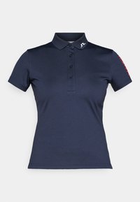 Jūras zila polo krekls, kas izgatavots no gludas auduma, ar apkakli, trīs pogu aizdari un sarkanu tekstu uz piedurknes. Logo uz apkakles.