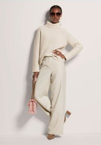 Beige gerippter Rollkragenpullover, cremfarbene weite Hose, hautfarbene High Heels und eine rosafarbene strukturierte Handtasche mit Kette. Große runde Sonnenbrille.