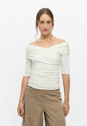 Femme à la peau claire et aux cheveux bruns porte un haut blanc à épaules dénudées froncé et un pantalon beige, debout devant un fond blanc uni.
