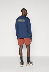 Marineblaues Langarmshirt mit buntem Logo auf dem Rücken, kombiniert mit orangefarbenen Shorts und schwarzen Socken, getragen mit mehrfarbigen Sportschuhen.