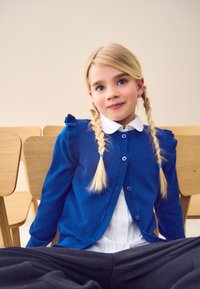 Cardigan blu lavorato a maglia con dettagli arricciati, chiusura con bottoni, indossato sopra una camicia bianca. Capelli biondo chiaro raccolti in trecce a due code, sfondo neutro.