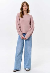 Hellrosa gerippter Pullover mit weiten Ärmeln, kombiniert mit hellblauen, hochgeschnittenen Schlagjeans und grauen Sneakers mit weißen Akzenten.