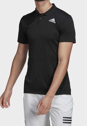 Homme portant un polo Adidas noir avec un logo sur la poitrine et un short blanc avec trois bandes noires sur la cuisse, sur fond uni.