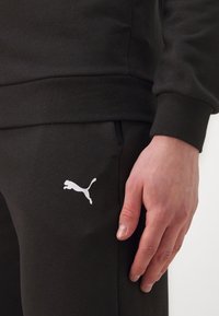 Felpa nera e pantaloni della tuta realizzati in tessuto morbido. Presentano un logo Puma bianco sulla gamba. I polsini e l'elastico in vita sono aderenti.