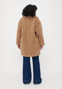 Casaco castanho, felpudo e oversized com detalhes na costura superior; possui mangas longas e bolsos laterais. Usado sobre jeans de boca de sino.
