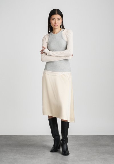 Top sin mangas gris ajustado de canalé debajo de una blusa de manga larga beige, combinado con una falda asimétrica de satén crema y botas altas negras.