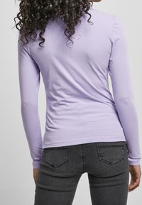 Dlouhý rukáv levandulový top vyrobený z měkké tkaniny, s přiléhavým designem a hladkým povrchem. V kombinaci s tmavě šedými džíny se zadními kapsami.