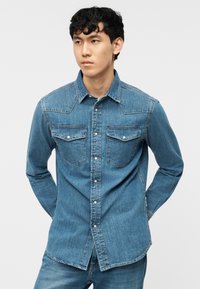 Νεαρός άνδρας όρθιος με τα χέρια πίσω από την πλάτη, φορώντας μπλε πουκάμισο τζιν με κουμπιά και δύο τσέπες στο στήθος, συνοδευόμενο από τζιν παντελόνι στο ίδιο χρώμα.