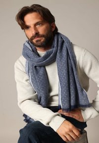 Écharpe bleue à motifs en tissu doux, drapée lâchement autour du cou. Présente un bord effiloché et est portée avec un pull léger en maille grise.