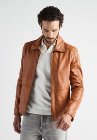 Brun læderjakke med lynlås og spidse kraver, med syninger og en glat overflade. Bæres over en lys sweater.