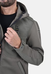 Giacca a hood da uomo in verde oliva, realizzata in tessuto liscio, con chiusura a zip, tasche a toppa e bottoni ai polsini.