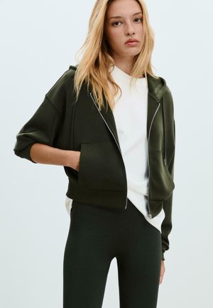 Žena s dlhými blond vlasmi, oblečená v tmavozelenej mikine na zips, ktorá má na sebe bielu košeľu a čierne legíny, stojaca s jednou rukou vo vrecku.