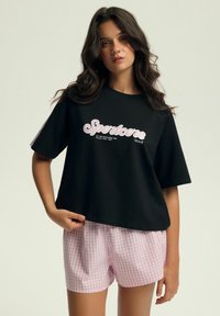 Czarny t-shirt z krótkim rękawem z napisem "Sportcore" w kolorze różowym, zestawiony z jasnoróżowymi szortami w biało-różową kratkę.