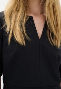 Černé šaty s véčkovým výstřihem, krátkými rukávy a hladkou texturou. Obsahují jemné boční kapsy a přiléhavý střih.