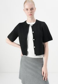 Black ribbed cardigan with short sleeves, round neck, and five white buttons, worn over a white top, paired with a grey skirt.