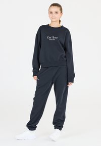Mörkblå sweatshirt med vit text "Les'Arcs" och matchande sweatpants, med avslappnad passform och muddar vid anklarna, parat med vita sneakers.
