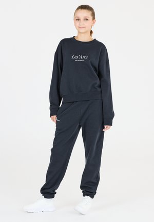 Mörkblå sweatshirt med vit text "Les'Arcs" och matchande sweatpants, med avslappnad passform och muddar vid anklarna, parat med vita sneakers.