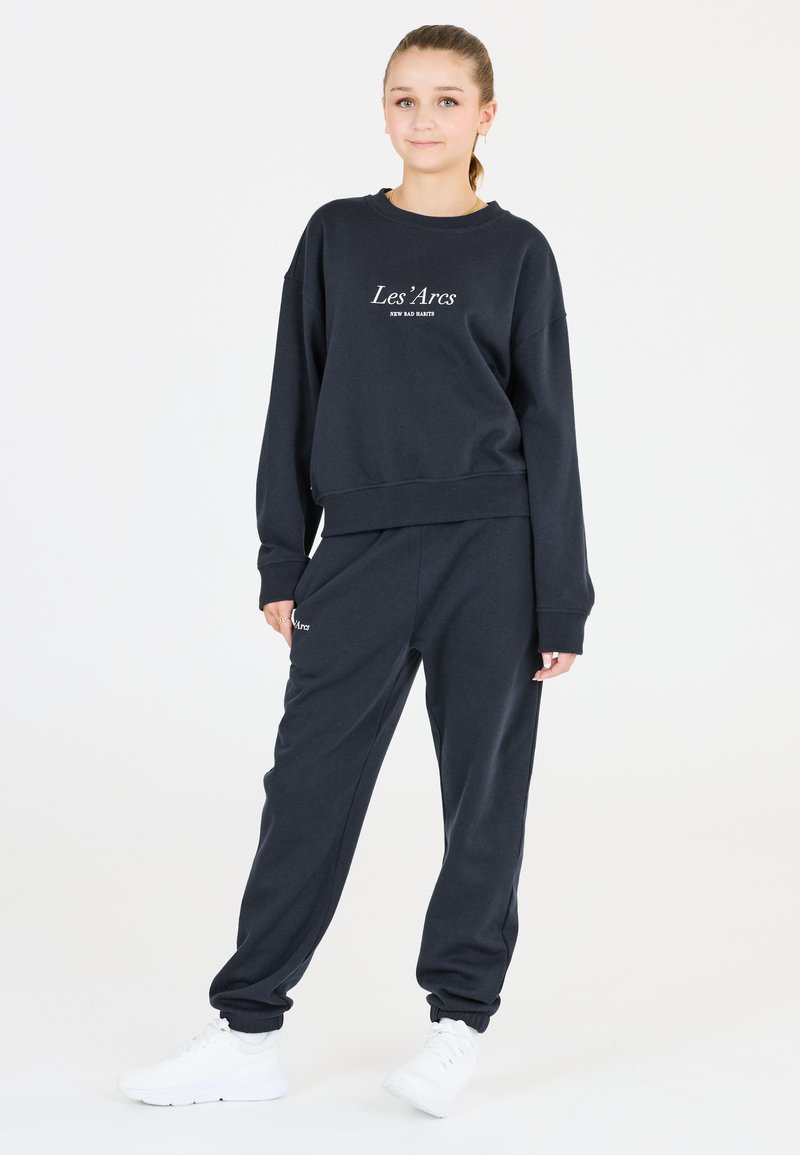 Mörkblå sweatshirt med vit text "Les'Arcs" och matchande sweatpants, med avslappnad passform och muddar vid anklarna, parat med vita sneakers.