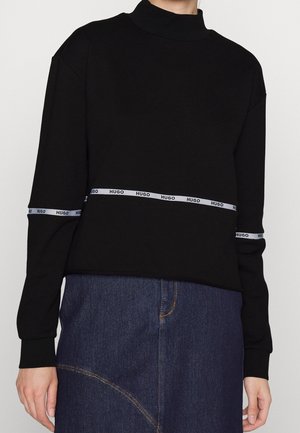 Zwarte top met lange mouwen met witte "HUGO" merkbanden rond de taille en onderarmen, gecombineerd met een donkerblauwe denimrok.