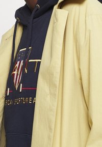 Żółta kurtka zewnętrzna na granatowym kapturze, z wyszywanym czerwono-złotym logo, zaciąganym sznurkiem i ściągaczami przy mankietach.