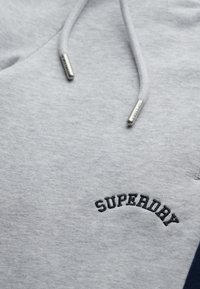 Lichtgrijze hoodie stof met zwart geborduurde "SUPERDRY" tekst en bijpassende trekkoorden met metalen uiteinden.