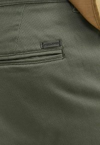 Tissu texturé vert avec un motif géométrique subtil, comportant une étiquette noire JACHS+JONES et un détail de poche dissimulée.
