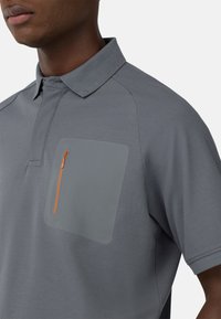Šedé polo tričko s límečkem, se velkou šedou náprsní kapsou a oranžovým zipem. Vyrobeno z hladké, strukturované látky.