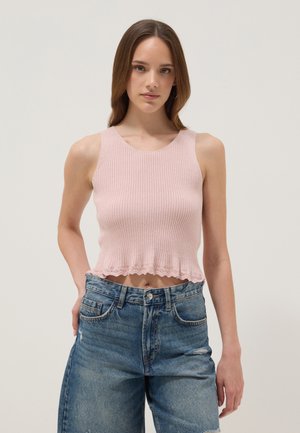 Jeune femme portant un crop top côtelé rose pâle sans manches et un jean en denim bleu taille haute sur un fond uni.
