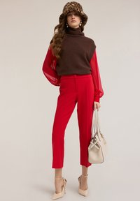 Brązowy sweter z dzianiny z golfem noszony pod czerwoną przezroczystą bluzką, do tego czerwone, dopasowane spodnie i beżowe buty na wysokim obcasie, trzymająca beżową torebkę.