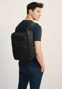 Czarny plecak z materiału z wyściełanymi szelkami, przednią kieszenią i eleganckim designem, noszony na plecach osoby w granatowej koszuli.
