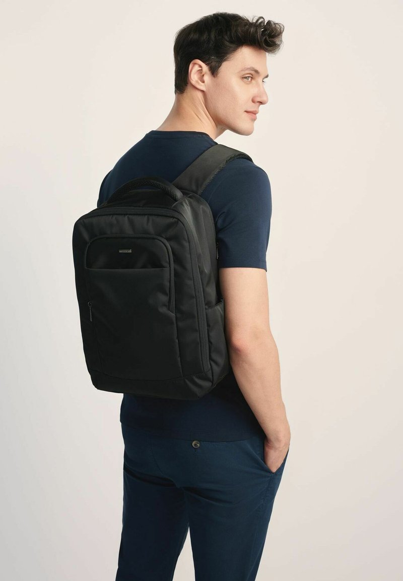 Czarny plecak z materiału z wyściełanymi szelkami, przednią kieszenią i eleganckim designem, noszony na plecach osoby w granatowej koszuli.