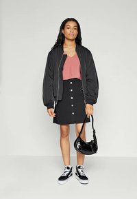 Zwarte bomberjack, lichtroze top, zwarte knopenrok en zwarte sneakers. Houdt een zwart leren schoudertas vast. Focus op casual stijl details.