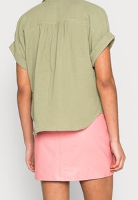 Chemise à manches courtes, légère, de couleur vert olive avec un pli dans le dos, associée à une mini-jupe en cuir rose brillante et ajustée.